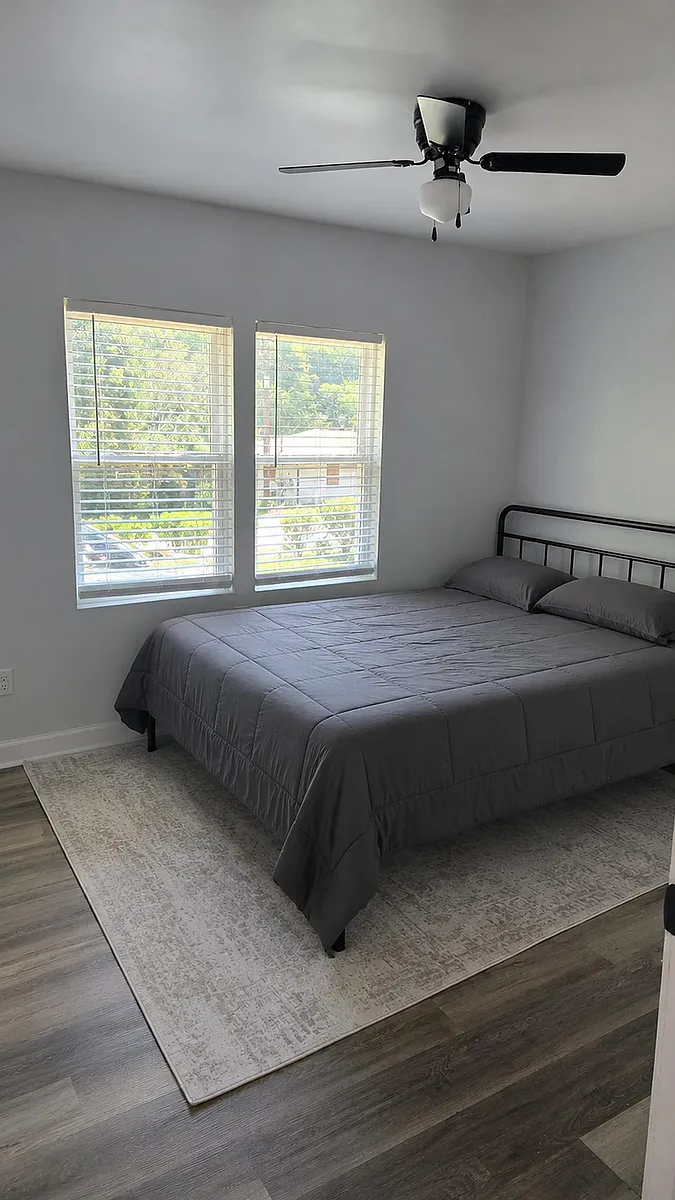 Photo 1 of 9 - Washington Rd - McDonough, GA Furnished bedroom with a queen bed, ceiling fan, and two windows with blinds for natural light and ventilation. - Washington Rd