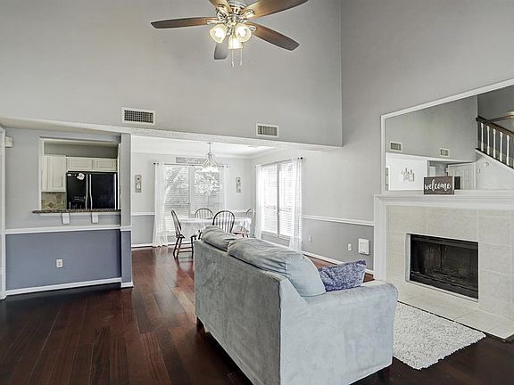 The living room features tall ceilings, dark wood floors, and a cozy fireplace. It is open to the dining room and kitchen.