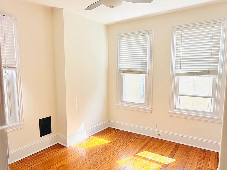 Bright corner bedroom with three windows on two walls, modern ceiling fan, and warm hardwood floors bathed in natural sunlight.