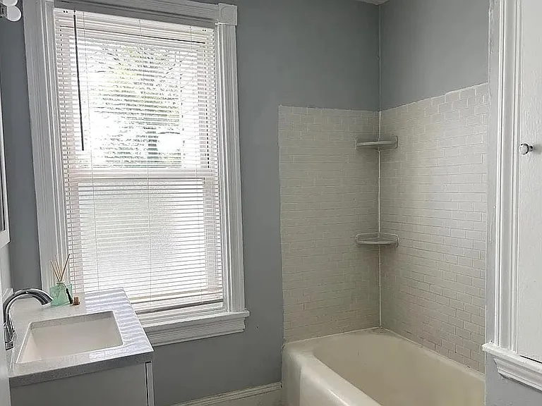 Updated bathroom with a full-size tub and shower, modern vanity, and classic tile surround. Bright window provides natural light, creating a clean and refreshing space.