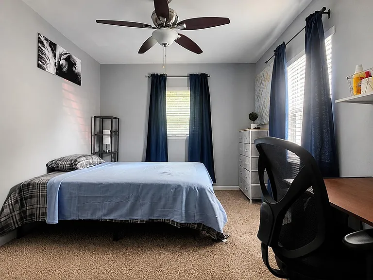 Furnished bedroom with ceiling fan, two windows with blinds, a work desk, and open wall shelving for extra storage.