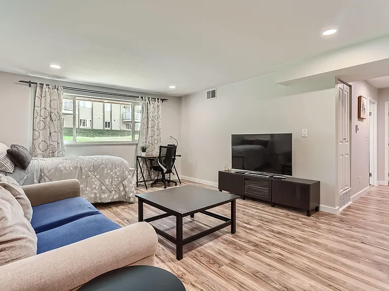 Furnished living area with sofa seating, TV console, and a work desk beside a large window with curtains.