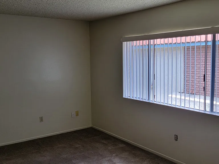 Carpeted bedroom with large window and vertical blinds at Magnolia Apartments in Riverside, California.