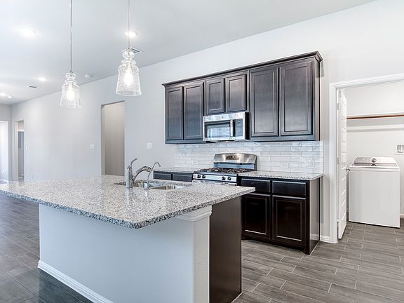 Kitchen island with sink, warm cabinetry, backsplash, and pendant lighting over island.