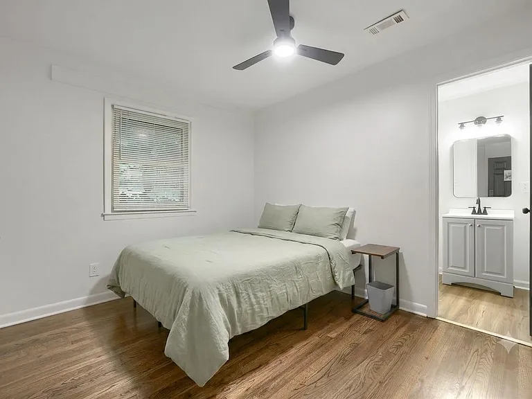 Spacious bedroom with hardwood floors, ceiling fan, and a window with blinds; convenient access to an adjacent bath.