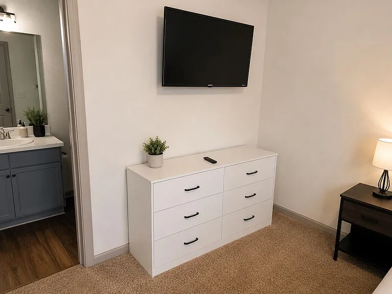 Bedroom features a six-drawer dresser and wall-mounted TV, with a bedside table and lamp for added convenience.