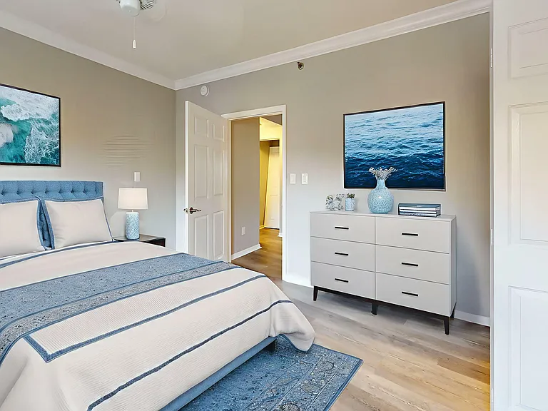 Elegant bedroom with coastal decor, featuring a plush blue headboard, soothing ocean artwork, and sleek dresser, creating a tranquil retreat.