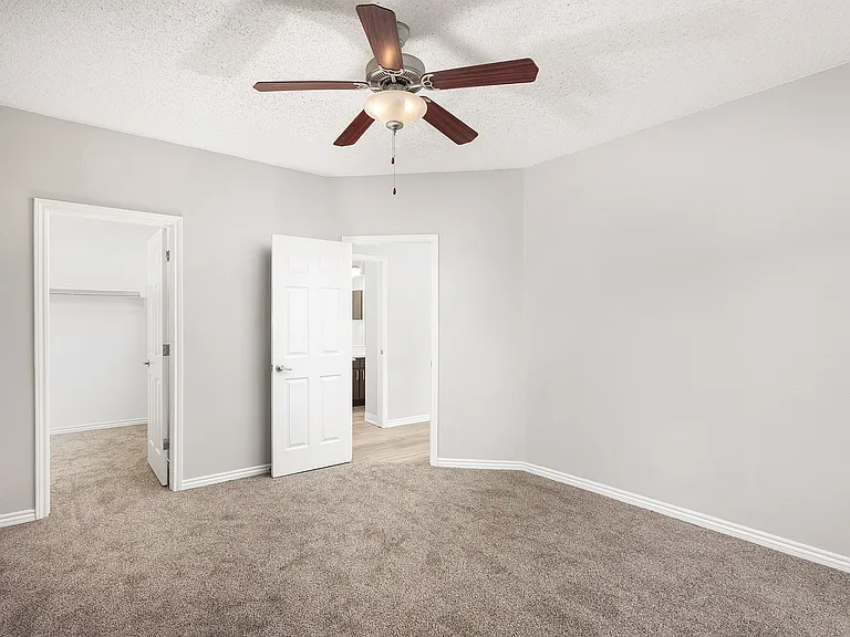 Bright and spacious bedroom with soft carpeting and modern ceiling fan, featuring white doors leading to a walk-in closet and a sunlit hallway.