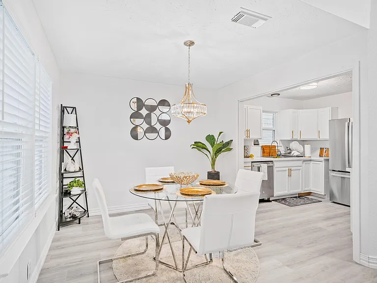 Glass-top dining area seats four under a chandelier, with easy flow into the adjacent kitchen and ample cabinet space.