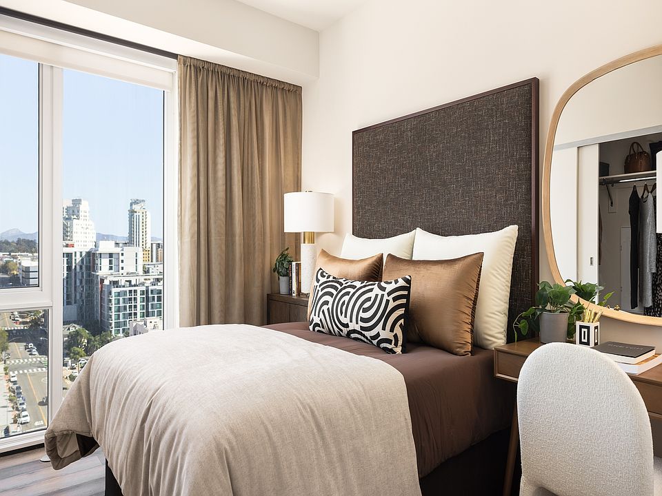 Chic and cozy bedroom featuring a plush bed with decorative pillows, elegant desk with mirror, and a stunning city view through large windows.