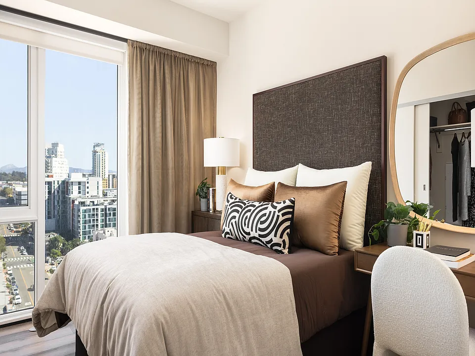 Chic and cozy bedroom featuring a plush bed with decorative pillows, elegant desk with mirror, and a stunning city view through large windows.