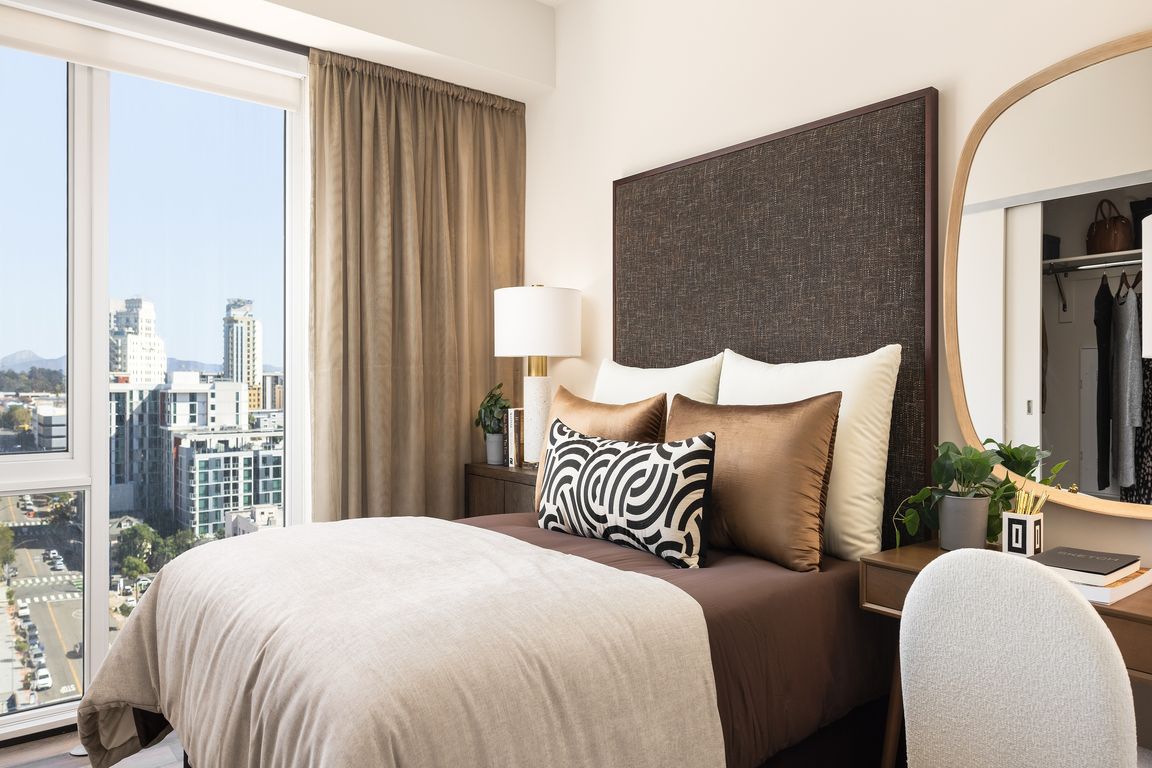 Chic and cozy bedroom featuring a plush bed with decorative pillows, elegant desk with mirror, and a stunning city view through large windows.