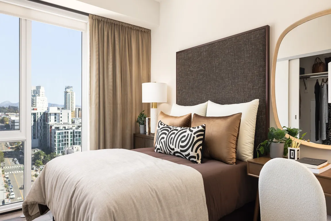 Chic and cozy bedroom featuring a plush bed with decorative pillows, elegant desk with mirror, and a stunning city view through large windows.