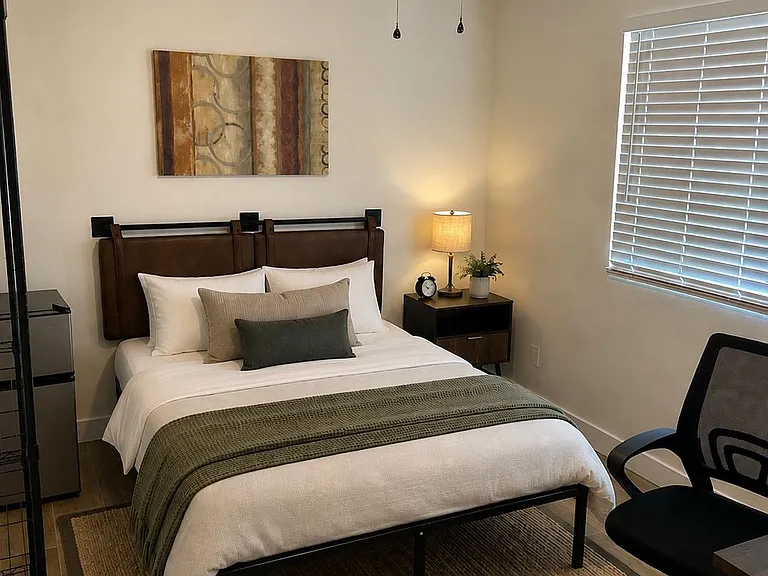 Furnished bedroom with a comfortable bed, bedside table and lamp, ceiling fan, and a window with blinds for privacy.