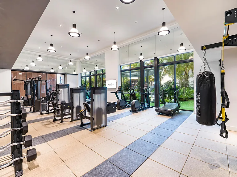 The fitness center at Modera Aventura features floor-to-ceiling glass, modern equipment, and views of the tropical courtyard.