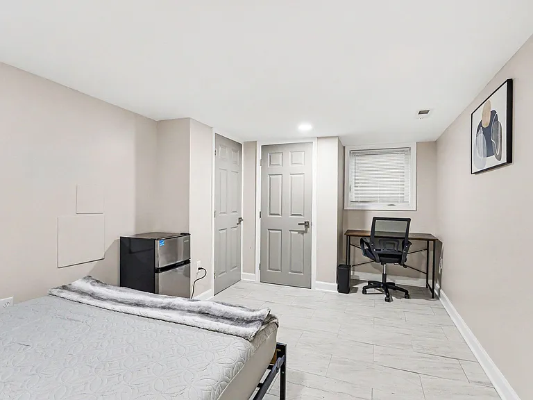 Private bedroom with a comfortable bed, in-room mini fridge, and a dedicated desk area beneath a window with blinds.