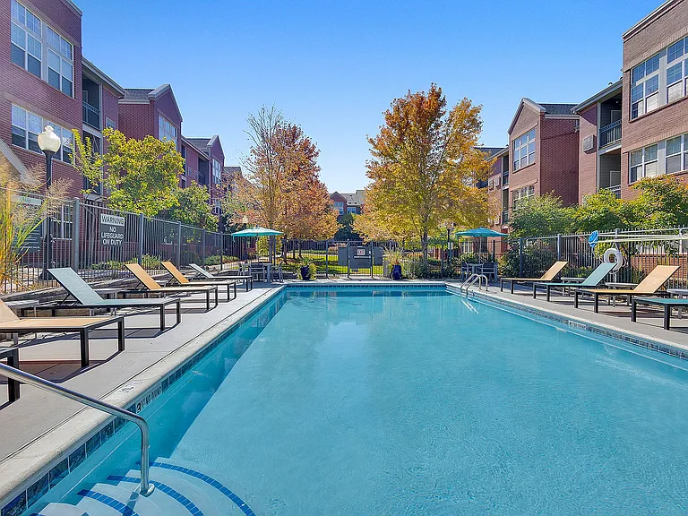 Inviting outdoor pool area with modern lounge chairs, surrounded by lush greenery and contemporary brick apartment buildings basking in sunlight.
