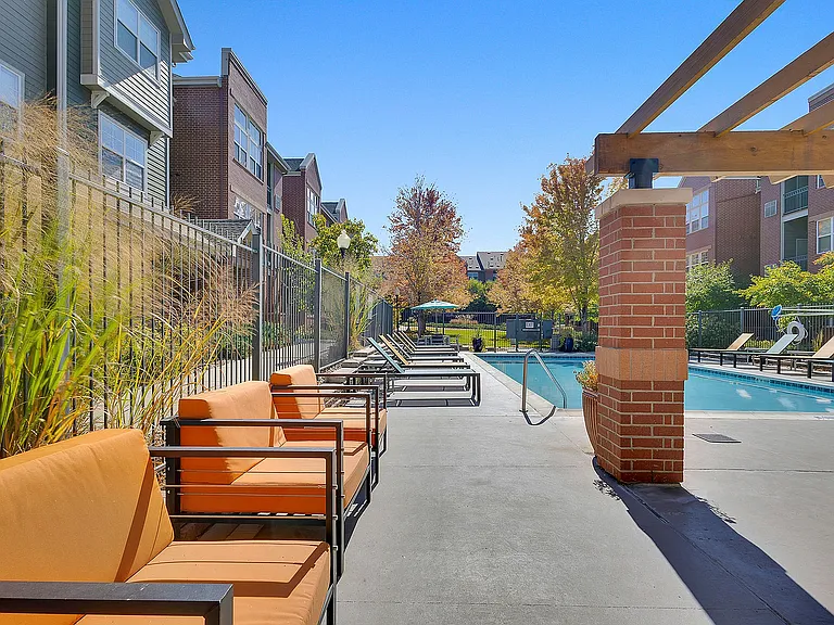Inviting outdoor pool area with stylish orange seating and sleek sun loungers, nestled beside modern apartment buildings and lush foliage.