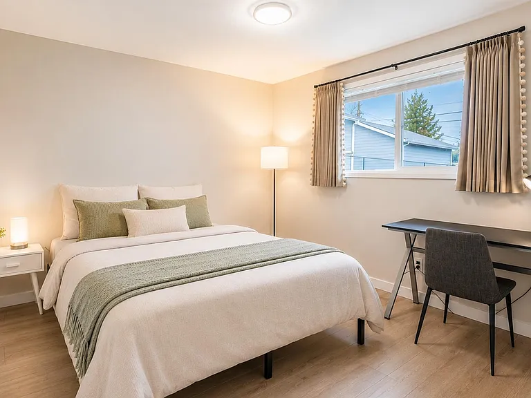 Well-sized bedroom with a furnished queen bed, nightstand, and dedicated desk workspace plus a large window with curtains.