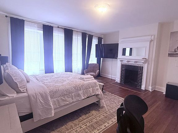 Spacious bedroom with hardwood floors, large windows, a mounted TV, and a decorative fireplace mantel for added character.
