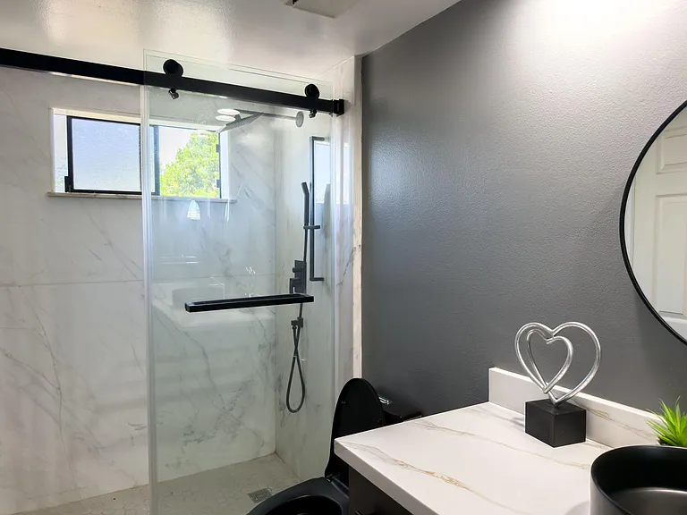 Walk-in shower with glass enclosure, large window, and marble-look tile paired with a spacious vanity and mirror.