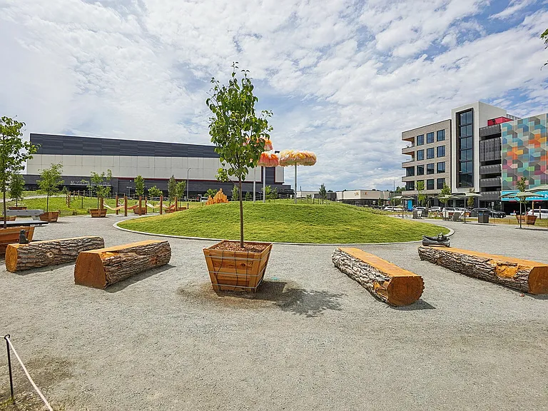 Vibrant urban park featuring lush green mounds, colorful benches, and modern art installations, nestled between contemporary buildings.