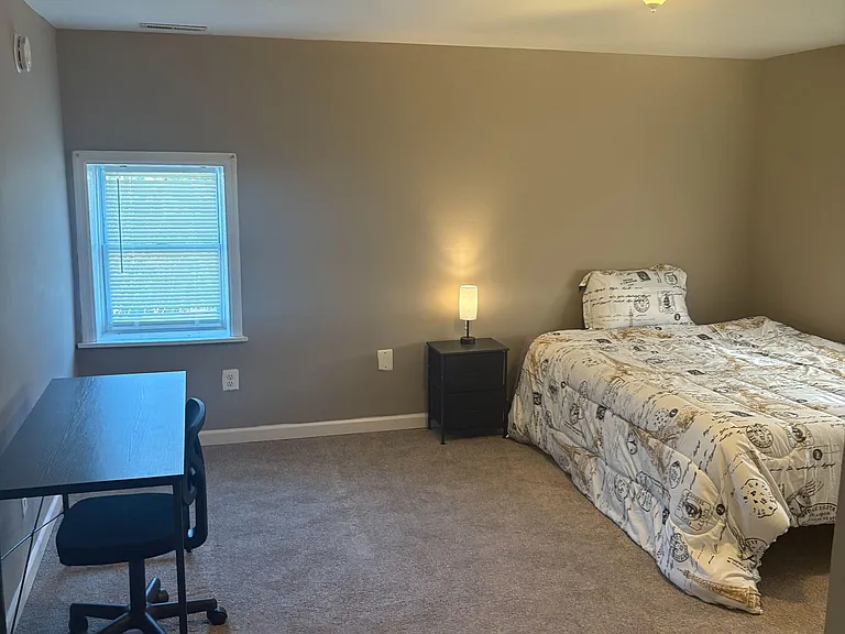 Furnished bedroom with a comfortable bed, nightstand lighting, and a dedicated desk area by the window.