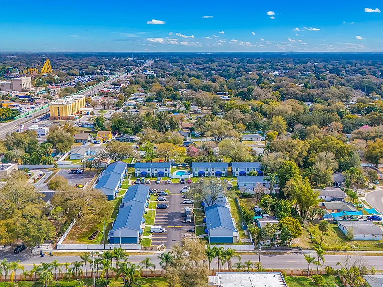Breathtaking aerial view of a serene suburban neighborhood, featuring a cluster of modern blue-roofed apartments and a central swimming pool, surrounded by lush greenery and vast landscapes under clear blue skies.