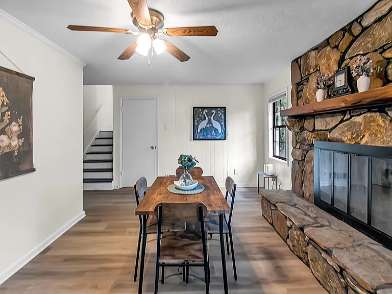 Dining area with a wood table, ceiling fan, and a stone fireplace mantle, plus nearby stair access and a side window.