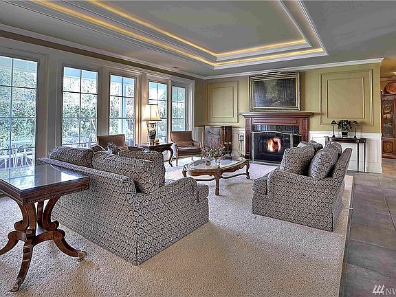 Living room offers gas fireplace with granite surround and custom mantle, wainscoting, tray ceiling with crown molding and accent lighting, wall of windows, and two sliders lead out to the covered patio. 