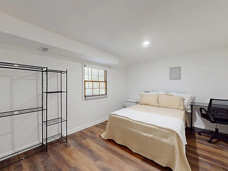Furnished bedroom with wood-look floors, a full bed, work desk and chair, open shelving, and a window with blinds.