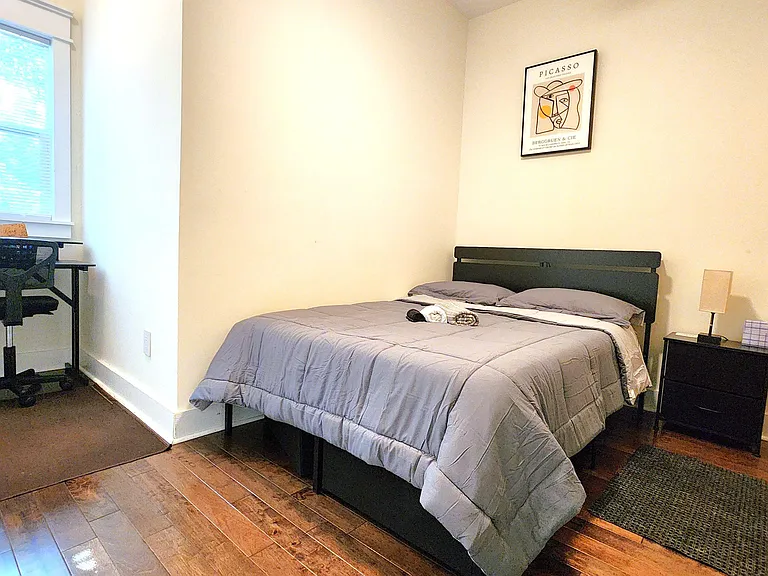 Furnished bedroom with a full bed, nightstand, and dedicated desk workspace, plus a window for natural light.