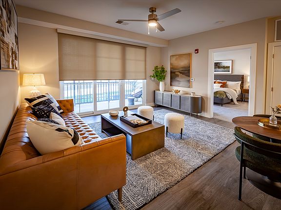 Elegant apartment living room with a chic leather sofa, modern decor, abundant natural light, and an open layout, including dining area, flowing into a cozy bedroom.