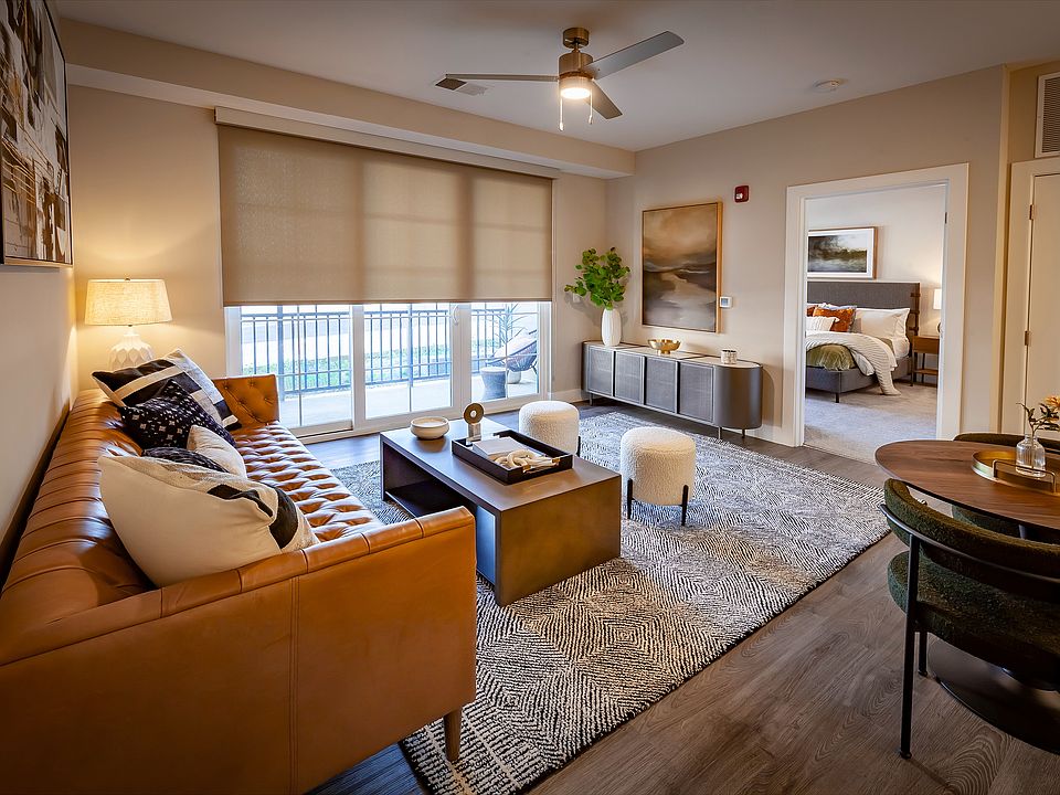 Elegant apartment living room with a chic leather sofa, modern decor, abundant natural light, and an open layout, including dining area, flowing into a cozy bedroom.