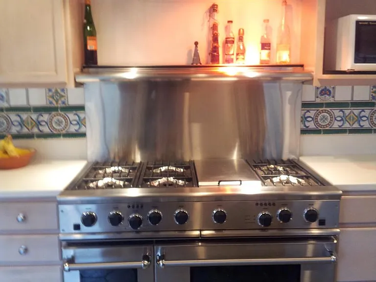 Commercial-grade stainless steel range with six gas burners, double ovens, and an overhead warming shelf with lighting.