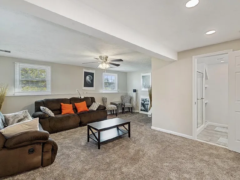 Spacious living room with plush carpeting, ceiling fan, and seating area, plus easy access to an adjacent full bath.