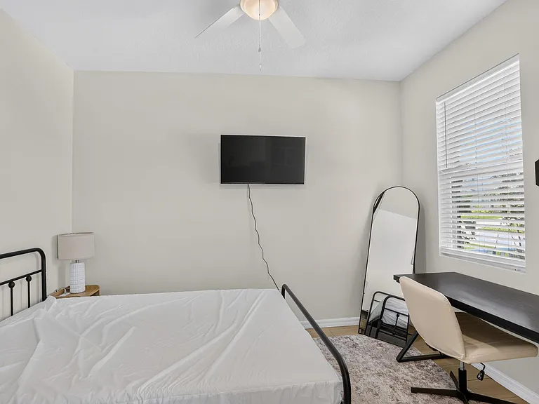 Private bedroom with a bed, wall-mounted TV, and dedicated desk setup, plus a large window with blinds for natural light.