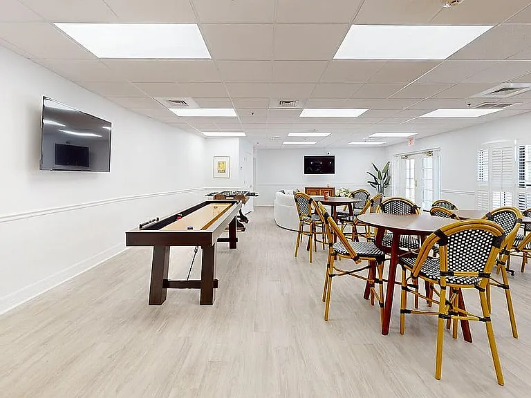 Bright and spacious recreational room featuring elegant seating, a shuffleboard table, and wall-mounted TV, perfect for social gatherings.