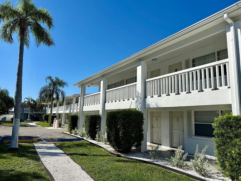 Charming two-story apartment building with white railings, surrounded by lush greenery and palm trees under a bright blue sky.