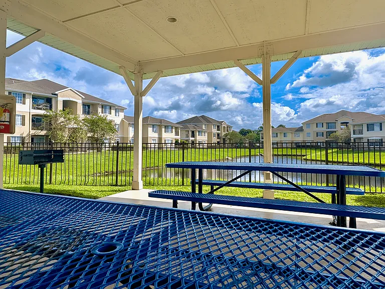 Serene apartment view from a covered patio with blue picnic tables, a barbecue grill, and lush greenery around a tranquil pond.