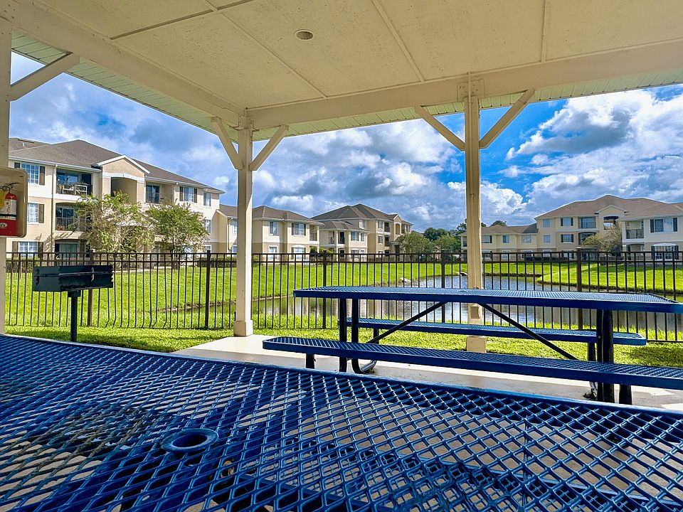 Serene apartment view from a covered patio with blue picnic tables, a barbecue grill, and lush greenery around a tranquil pond.