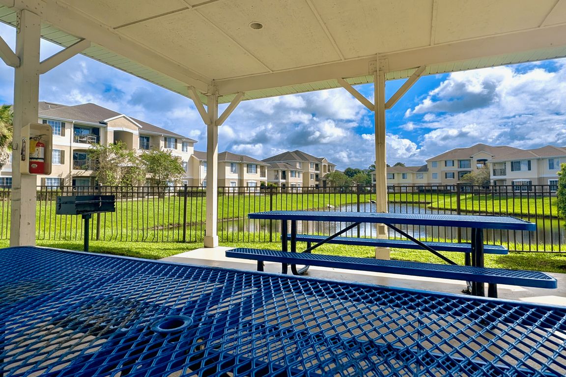 Serene apartment view from a covered patio with blue picnic tables, a barbecue grill, and lush greenery around a tranquil pond.