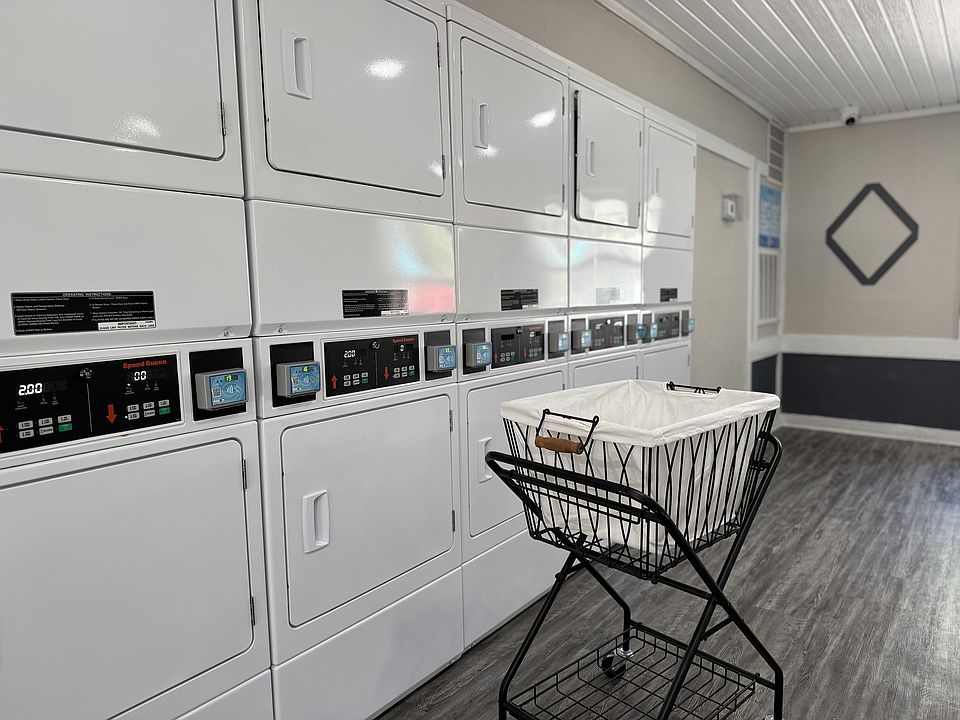 Modern apartment laundry room featuring a sleek row of white washers and dryers with a stylish laundry cart on polished wood flooring.
