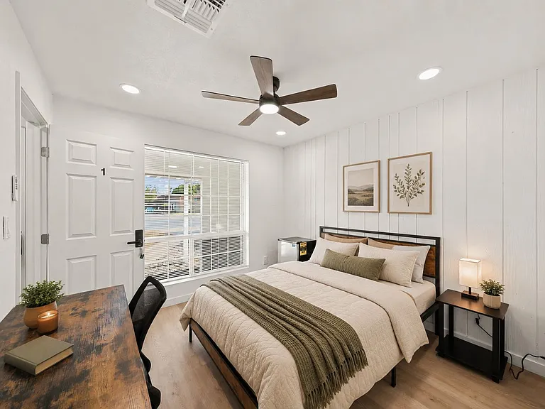 Well-appointed bedroom with a full bed, work desk, ceiling fan, and a large window with blinds for natural light.