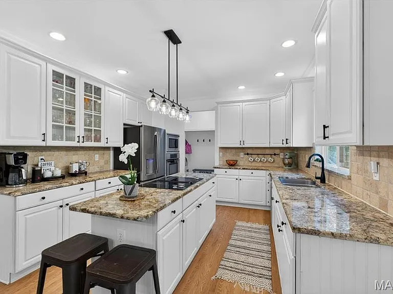 Bright kitchen featuring a central island with a built-in cooktop, light wood-finish flooring, white cabinetry with black hardware, granite countertops, and a tiled backsplash