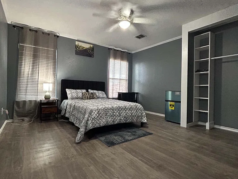 Spacious furnished bedroom with two windows, ceiling fan, wood-look flooring, and open shelving plus a mini fridge.