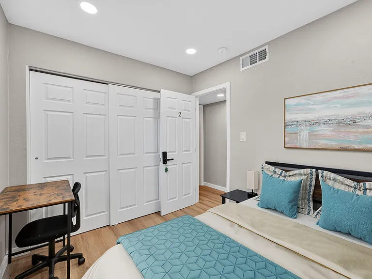 Furnished bedroom with a full bed, nightstand lighting, a dedicated desk workspace, and a spacious sliding-door closet.