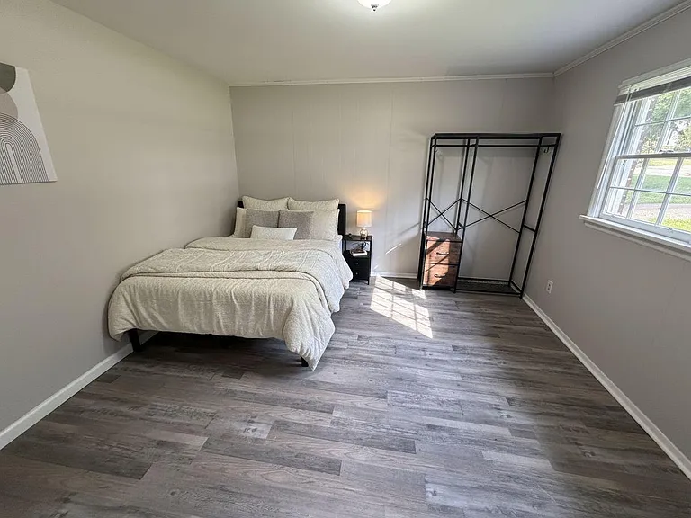 Spacious bedroom with wood-look flooring, large window for natural light, bedside table, and open closet storage.
