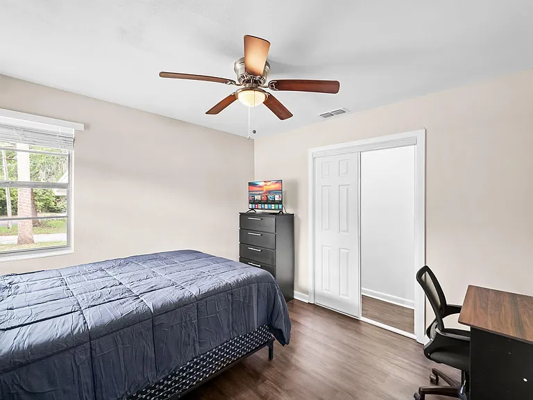 Furnished bedroom with a full bed, dresser with TV, dedicated desk space, ceiling fan, and a sliding-door closet.