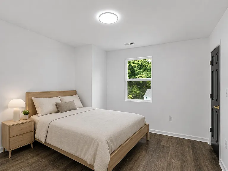 Spacious bedroom with wood-style flooring, a large window with leafy views, and ample wall space for storage or a desk.